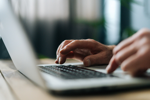 Close-up side view of unrecognizable mature adult man writing text on portable wireless computer sitting at desk in dark living room. Unknown senior male fingers typing laptop, selective focus.