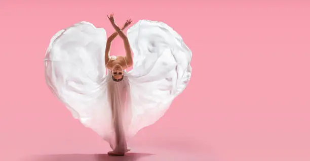 slender graceful ballerina in pointe shoes waves a long white skirt showing a heart shape on a pink background