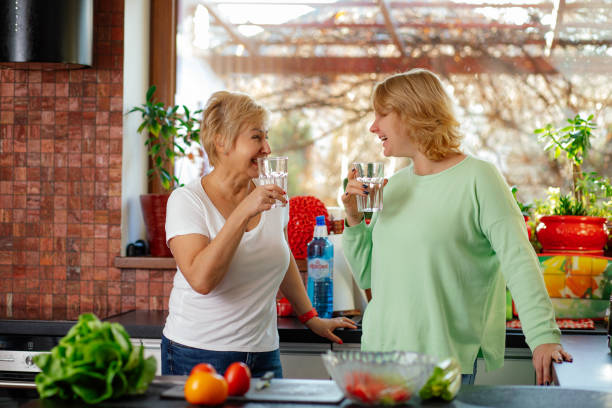 la mère et la fille blondes et souriantes boivent de l’eau et parlent à la cuisine lumineuse avec une grande fenêtre. la famille passe du temps ensemble - water women glass healthy eating photos et images de collection