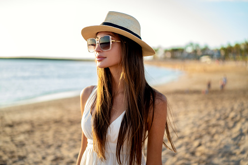 Young beautiful woman enjoying summer vacation, walking at the beach. Portrait of brunette woman with long hair wearing fashionable sunglasses and hat. Tourism. Real people lifestyle.