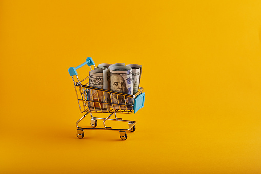 Shopping cart with rolled American One Hundred Dollar Bills on yellow / orange background