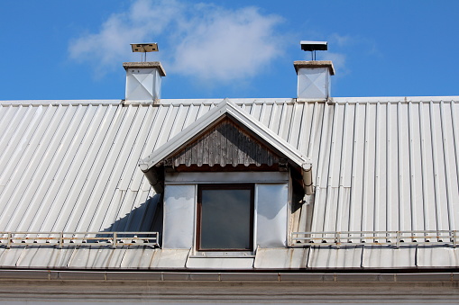 Old roof window with wooden frame and wooden dilapidated decorative boards on top covered with newly installed metal roof tiles surrounded with snow guards and two metal sheets covered chimneys on top of suburban family house on clear blue sky background