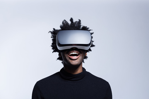 Portrait of excited afro american man wearing virtual reality glasses, standing against grey background and laughing.