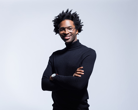 Friendly afro american young man wearing black turtleneck and glasses, standing with arms crossed and smiling at camera. Studio shot on grey background.