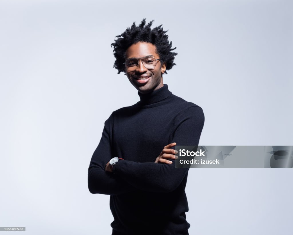 Portrait of confident man in black turtleneck Friendly afro american young man wearing black turtleneck and glasses, standing with arms crossed and smiling at camera. Studio shot on grey background. Portrait Stock Photo