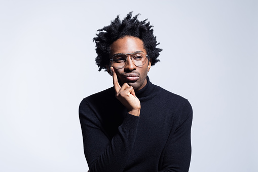 Handsome afro american young man wearing black turtleneck and eyeglasses, looking away with hand on chin. Studio shot on grey background.