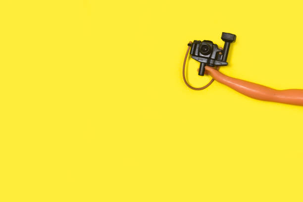 A doll hand holding a photo camera on a yellow background with copy space
