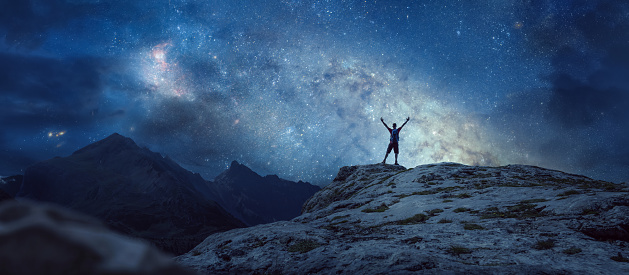 A silhouette of a hiker with raised arms under a beautiful starry sky. Panoramic image with copy space.