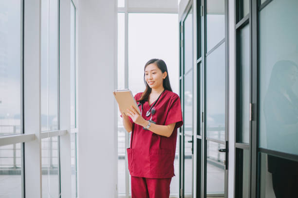 chinese female nurse healthcare taker smiling looking at clipboard on patient's health report in hospital corridor chinese female nurse healthcare taker smiling looking at clipboard on patient's health report in hospital corridor red medical scrubs stock pictures, royalty-free photos & images