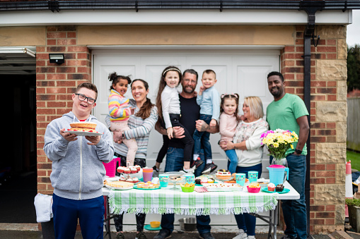 Family having a bake sale in their garden in the North East of England. They have hand made foods on a table ready to sell. One of the sellers has down syndrome and is standing holding a cake he has made. They are looking at the camera standing together.