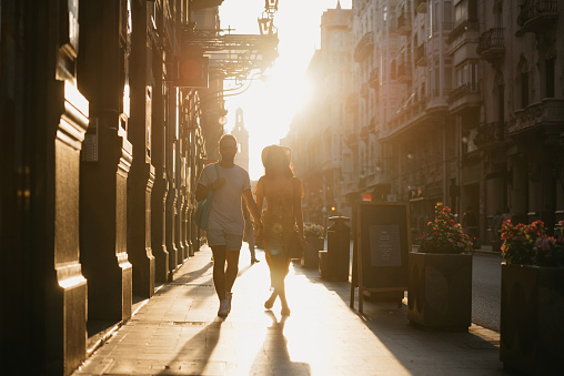 A girl in a hat and a yellow dress with a plunging neckline and her boyfriend with a beard are walking holding each other's hand in Spain. A couple of lovers on a date on the sunset in Valencia.