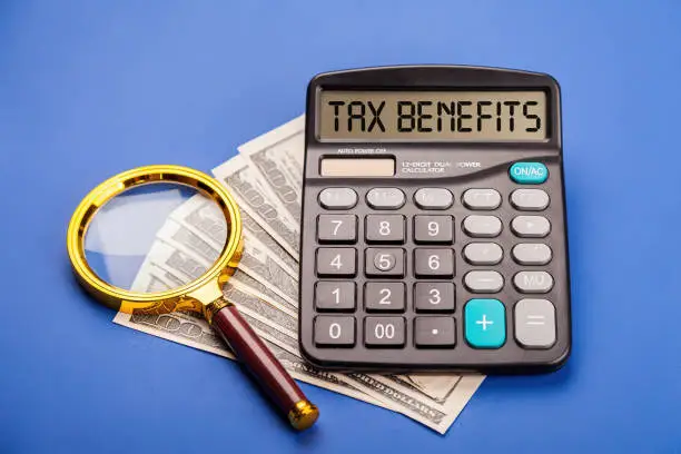 Photo of TAX BENEFITS. The concept of finance and business. On the blue background lies money dollars magnifying glass and a calculator with the inscription - TAX BENEFITS