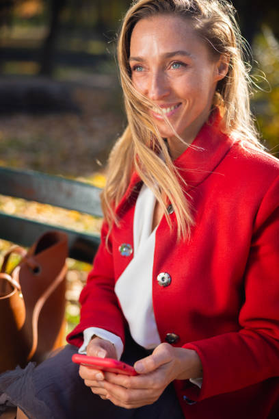 Elegant woman sitting on park bench on sunny autumn day and smiling, using smart phone for electronic banking, finances on the move