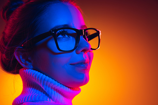 Closeup young beautiful girl in eyewear isolated on orange background in neon light, filter. Concept of emotions