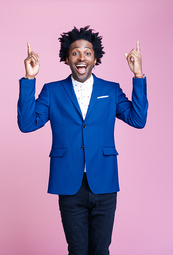 Cheerful afro american man wearing navy blue jacket and white shirt, pointing with index fingers at copy space, looking at camera. Studio shot on pink background. Portrait of designer.