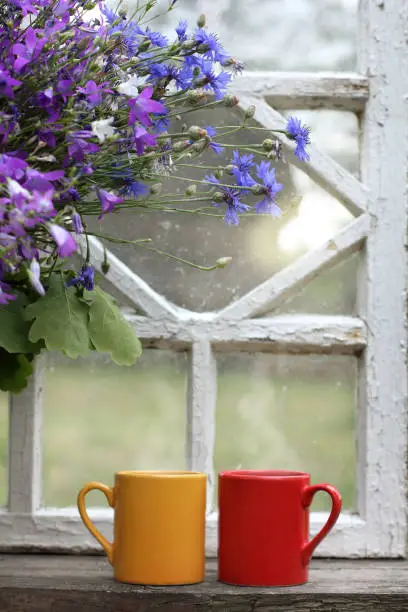red and yellow mugs next to bouquet of wild flowers on the windowsill against the backdrop of retro window