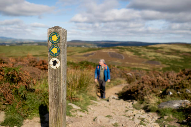 Out of focus shot of a senior man wearing a raincoat hiking uphill in Rothbury, Northumberland. The trail post is in the forefront of the shot.