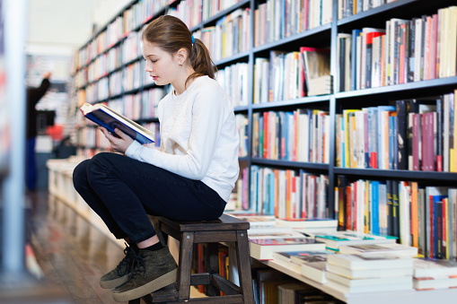 Cute little glad   positive girl absorbed in reading book while sitting on small wooden ladder in bookstore