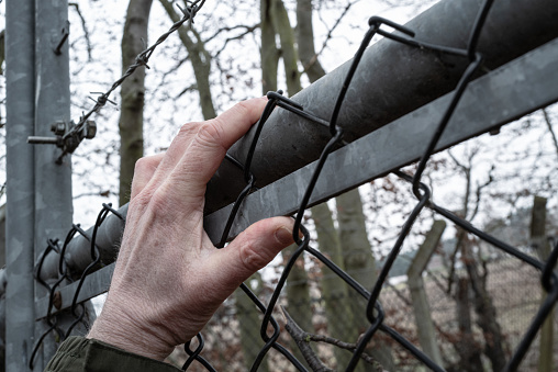 Protester seen climbing a containment fence with barbed wire. Trying to get into a fenced off, secure complex. His cold hand is gripping a allow fence beam.