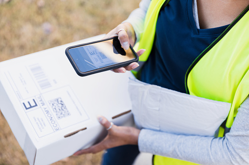 A high angle view of a female delivery person as she uses a smart phone to scan the label on the white cardboard box.  Both the label and the smart phone screen are visible.
