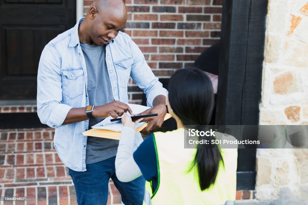 Female delivery driver holds tablet for customer to sign The unrecognizable young adult female delivery driver holds out a digital tablet for the mid adult male customer to sign.  They are standing on the front doorstep of the home. Delivering Stock Photo