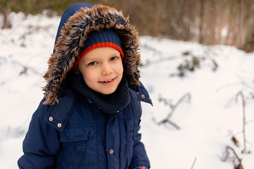Photo of a little boy enjoys winter activities that snow brings. Portrait of a cute boy wearing a warm jacket with cap on a winter day. Happy little boy enjoying show. Cute toddler enjoying first snow