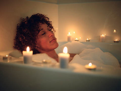 A woman relaxing in a bathtub, full of bubbles and surrounded by candles.
