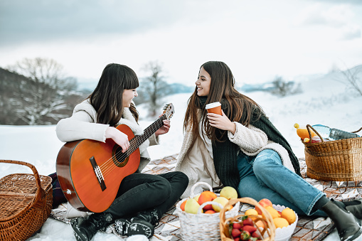 Two Cute Females Enjoying Guitar Music With Coffee And Snacks During Winter Mountain Picnic