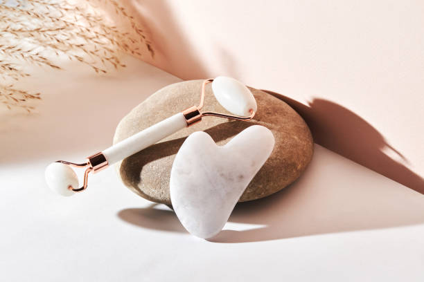 White quartz facial massage Jade roller, Gua Sha, on a river pebble stone on a beige background. stock photo