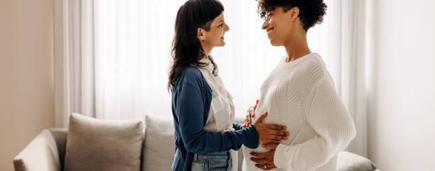 Smiling woman feeling a pregnant woman's belly Smiling woman feeling a pregnant woman's belly. Happy young woman feeling the movement of a pregnant woman's baby. Cheerful young woman spending time with her surrogate at home. surrogacy stock pictures, royalty-free photos & images