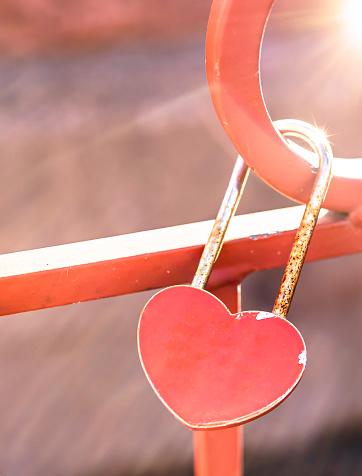 Rusty padlock of heart shape hold at red bridge with blurred background and bright sunlight. Traditional symbol for strengthen, loyalty, forever mutuality and eternity love. Romantic Valentine concept