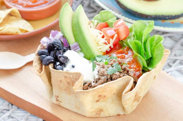 beef taco salad prepared with salad,avocado, tomato, salsa, ground beef, black beans, cheddar cheese, red onion and topped with sour cream and cilantro in a homemade baked flour tortilla bowl served on a wooden kitchen board in a mexican-style salad