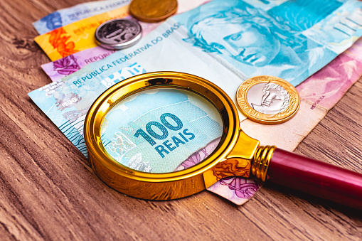 Billetes reales brasileños con una lupa en ellos y algunas monedas que están encima de un mueble de madera. Época de crisis financiera con alta inflación. photo