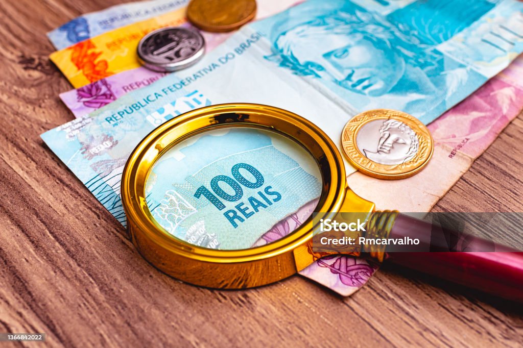 Brazilian Real banknotes with a magnifying glass on them and some coins that are on top of a wooden furniture. Time of financial crisis with high inflation. Tax Stock Photo