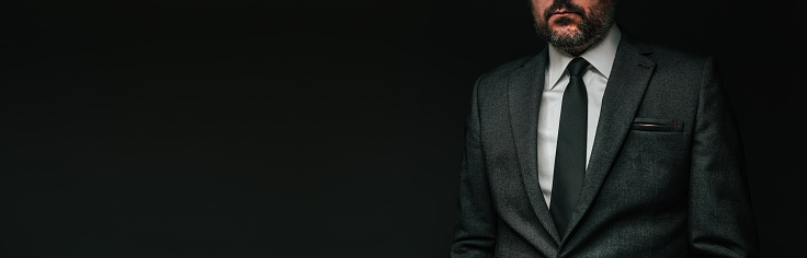 Panoramic low key portrait of handsome confident businessman in elegant grey suit posing against dark background
