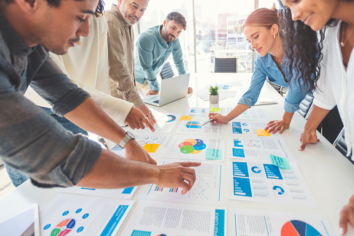 Multi racial group of people working with Paperwork on a board room table at a business presentation or seminar. The documents have financial or marketing figures, graphs and charts on them. People are pointing to different documents. There is a laptop on the table