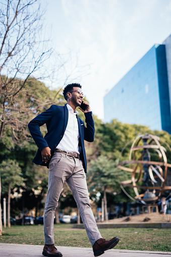 Elegante hombre de negocios negro sonriente caminando en un parque mientras usa su teléfono inteligente photo