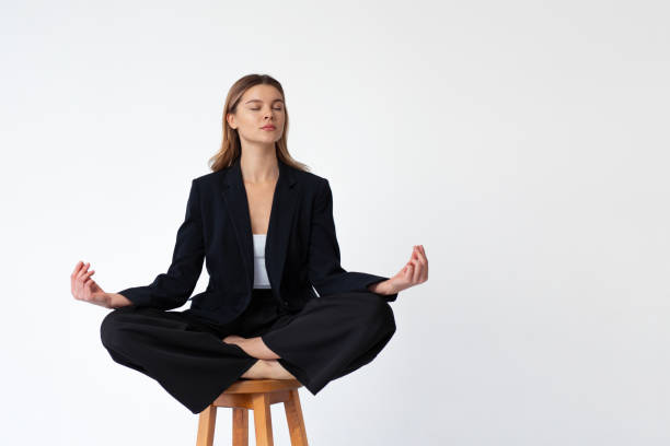 Young fashionable business woman sitting high bar wooden chair lotus pose dressed black office suit isolated in white background Young fashionable business woman sitting high bar wooden chair lotus position closed eyes dressed black office suit isolated in white background. Caucasian female business person meditation concept woman sitting cross legged in chair stock pictures, royalty-free photos & images