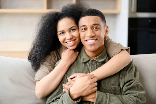 istock Smiling woman hugging from behind her handsome man, while looking at the camera. Young multiracial couple spending time in cozy modern living room 1366859413