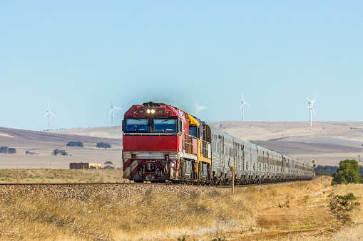 Overland Passenger Train passing wind turbines in outback Australia. Red and yellow locomotives leading long line of stainless steel passenger carriages with rolling hills and wind turbines in distance. Telephoto perspective. ID & logos edited