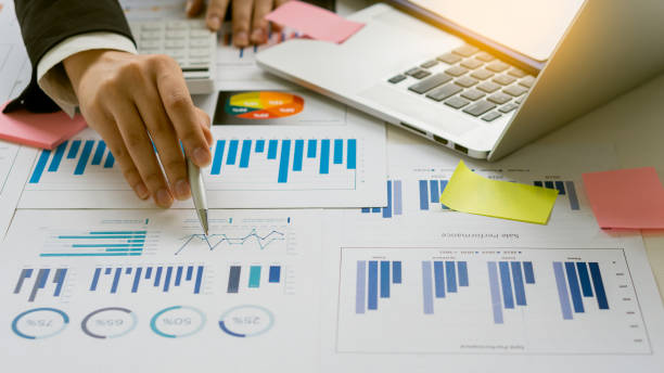 istock Businessman holding a profitability statistics analysis pen from a graph document in hand with a calculator and laptop on the table to plan profit growth from the past year. 1366894147