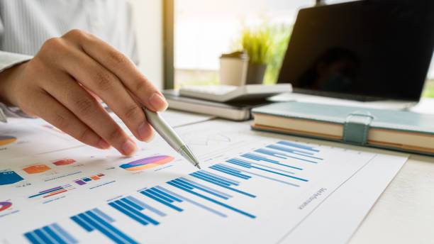 Businessman holding a profitability statistics analysis pen from a graph document in hand with a calculator and laptop on the table to plan profit growth from the past year. empresario sosteniendo un bolígrafo de análisis de estadísticas de rentabilidad de un documento gráfico en la mano con una calculadora y una computadora portátil sobre la mesa para planificar el crecimiento de las ganancias del año pasado.