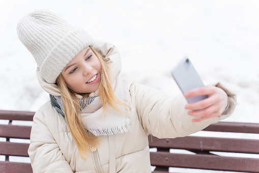cute caucasian girl in a knitted hat communicates by video communication while sitting on a bench in a winter park
