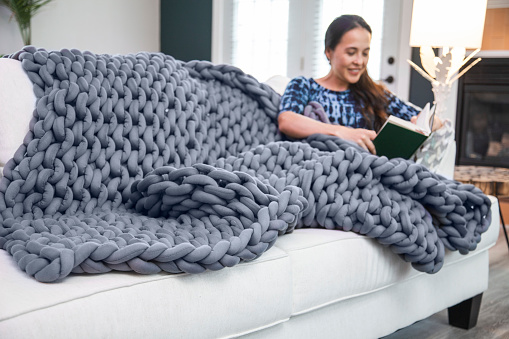 A woman reads and uses a gray knitted style weighted blanket in a bright modern living room on a white couch