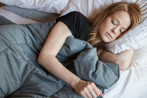 Young beautiful redhead woman sleeping while using soft weighted blanket in her beige eclectic apartment