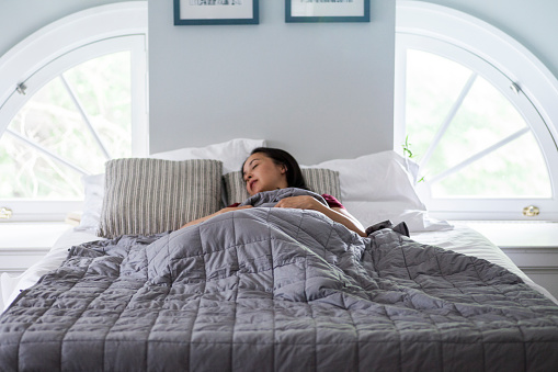A woman sleeps on a white bed with a soft weighted blanket in a bright open airy bedroom