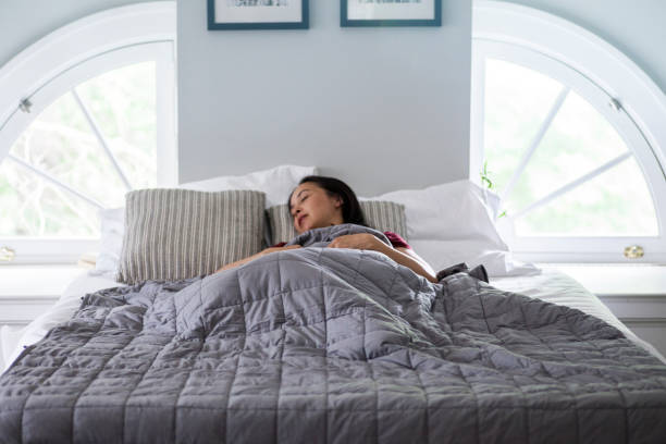 Woman sleeps under weighted blanket in bright bedroom A woman sleeps on a white bed with a soft weighted blanket in a bright open airy bedroom blanket stock pictures, royalty-free photos & images