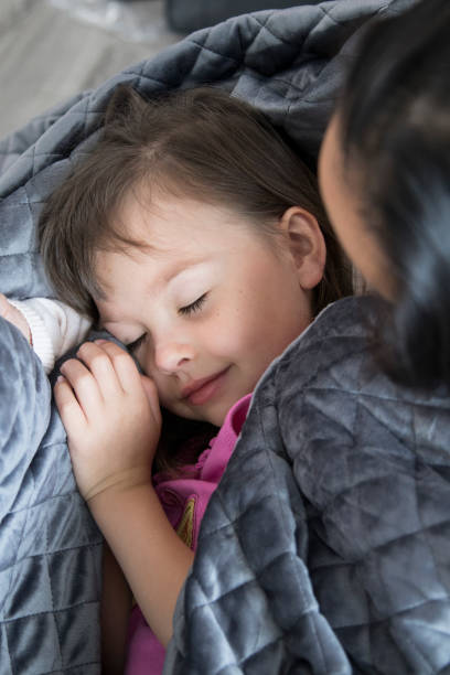 Mother tucks daughter into bed with weighted blanket stock photo