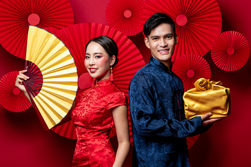 Smiling Asian couple in traditional costumes with fan and gold package as a gift for Chinese new year in oriental red background decoration