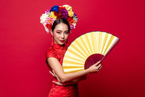 Beautiful Asian woman wearing traditional dress with flower chaplet holding golden fan in isolated studio red background for Chinese new year concept